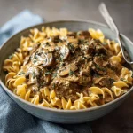 Close-up of a bowl of creamy beef stroganoff served over egg noodles, garnished with parsley.