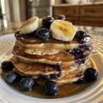 Close-up of fluffy blueberry banana pancakes stacked on a plate, topped with fresh blueberries and syrup.