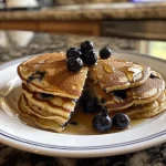 Close-up side view of fluffy blueberry pancakes stacked on a plate, topped with fresh blueberries.