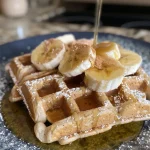 ['Close-up view of golden-brown Flourless Banana Oat Waffles topped with slices of banana and a sprinkle of cinnamon.', 'Side view of fluffy Flourless Banana Oat Waffles drizzled with syrup and garnished with fresh fruit.', 'Delicious Flourless Banana Oat Waffles stacked on a plate, featuring a textured surface and vibrant banana slices.', 'Moist Flourless Banana Oat Waffles displayed with a light dusting of cinnamon and almond milk on the side.']