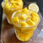['Close-up view of a glass of Tropical Mango Lemonade with mango and pineapple chunks.', 'Refreshing Tropical Mango Lemonade served in a clear glass, highlighted by vibrant yellow colors.', 'Side view of a chilled glass of Tropical Mango Lemonade garnished with lemon slices.', 'A refreshing glass of Tropical Mango Lemonade with ice, showcasing its bright color and fruit pieces.']