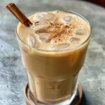 ['Close-up view of a refreshing iced chai latte in a clear glass with ice cubes.', 'Iced chai latte sitting on a wooden table, showcasing its creamy texture and spices on top.', 'Side view of a chilled iced chai latte garnished with chai spices, placed beside a colorful cloth.', 'Iced chai latte in a glass with brown tones, showing ice and layered milk.']