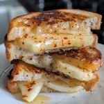 ['Close-up shot of a grilled cheese sandwich with melted cheddar, apple slices, and hot honey drizzle.', 'Side view of a Sweet and Spicy Hot Honey Apple Cheddar Grilled Cheese on whole grain bread.', 'Juicy grilled cheese sandwich showcasing sharp cheddar, apple slices, and honey drizzle.', 'A mouthwatering grilled cheese with apple slices and spicy honey, set on textured whole grain bread.']